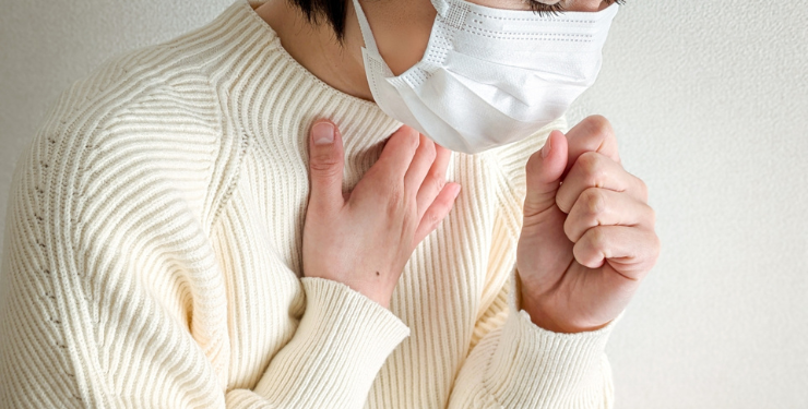 白いセーターを来た女性がマスクを付けて咳をしている様子